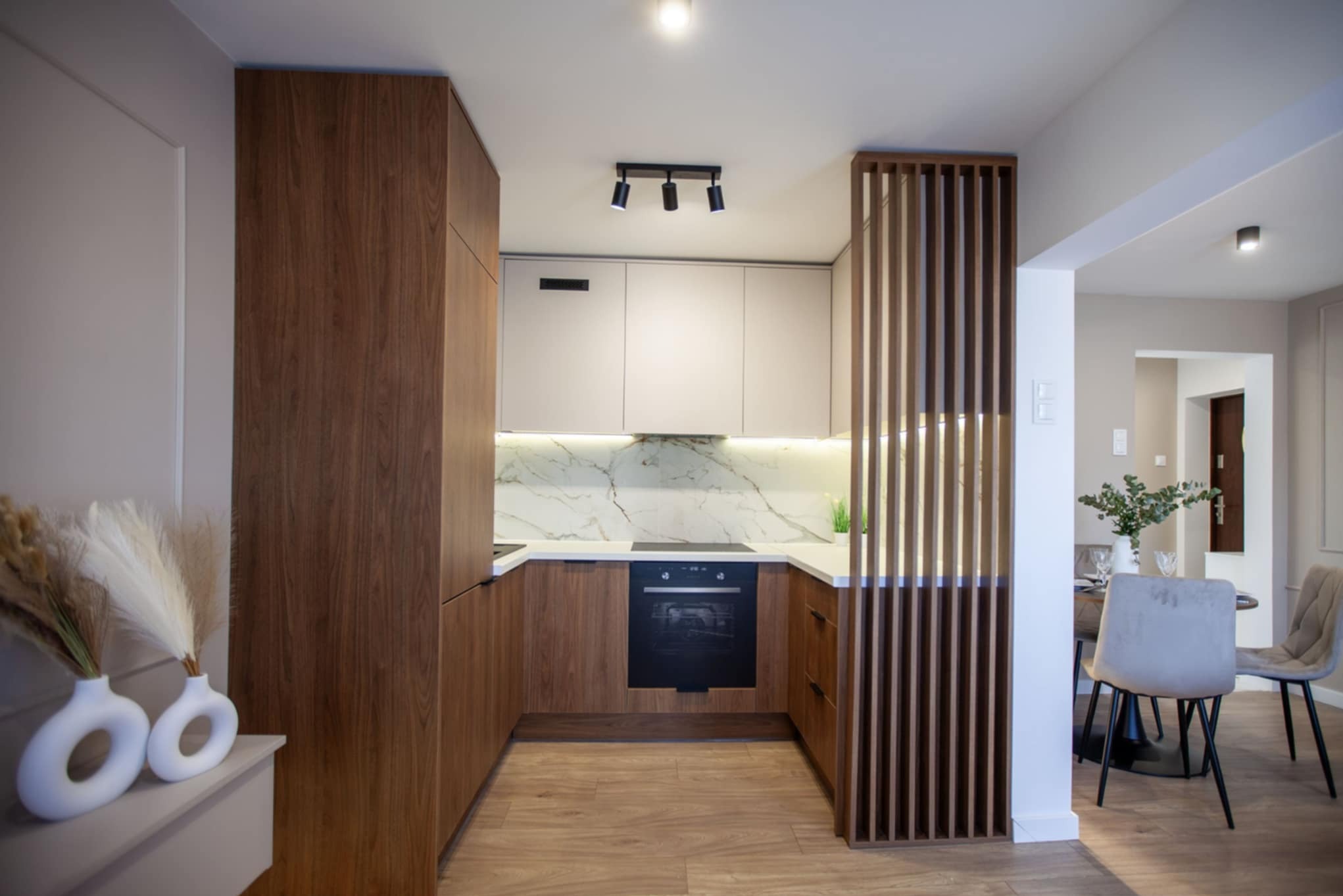 Nowoczesna kuchnia z drewnianymi szafkami, białym blatem i marmurowym backsplashiem, oddzielona drewnianą ażurową ścianką od jadalni z okrągłym stołem.