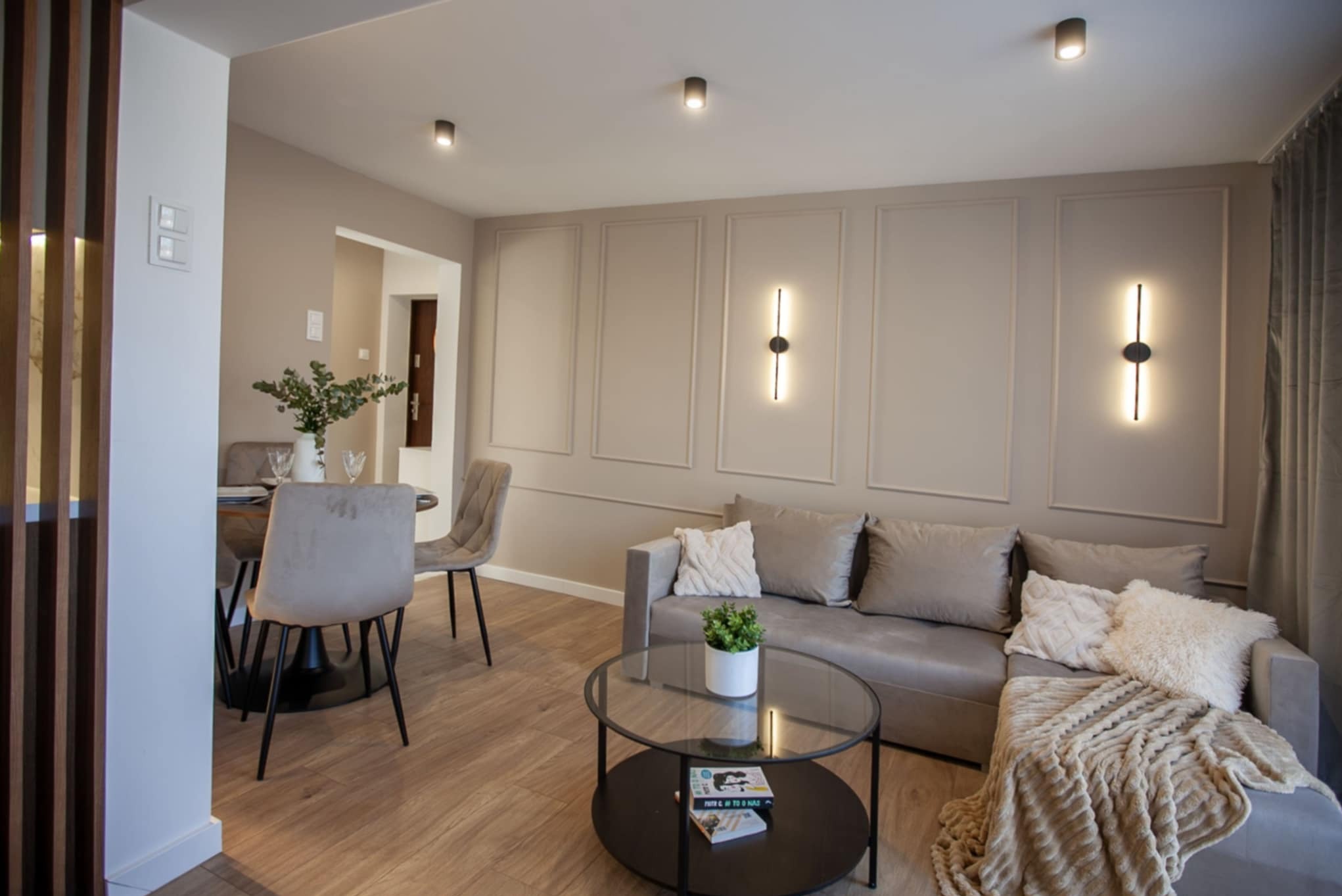 Neutralna kolorystyka salonu z drewnianą podłogą, szarą sofą z dekoracyjnymi poduszkami, szklanym stolikiem kawowym z książkami, okrągłym stołem z krzesłami i roślinami w wazonie, ozdobnymi...