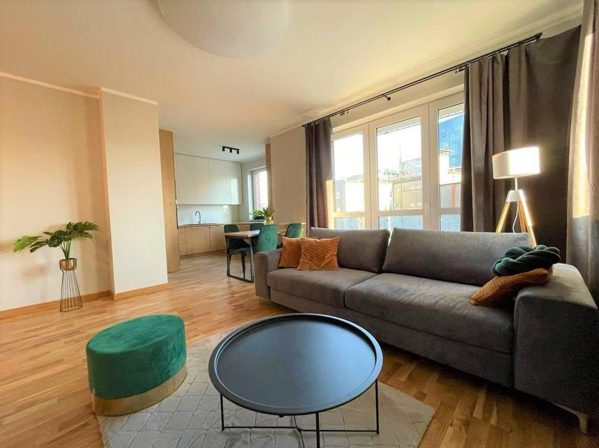 Jasne wnętrze mieszkania po remoncie: parkiet, szara sofa z poduszkami, okno z zasłonami, okrągły stolik kawowy i widok na kuchnię z jadalnią w tle.