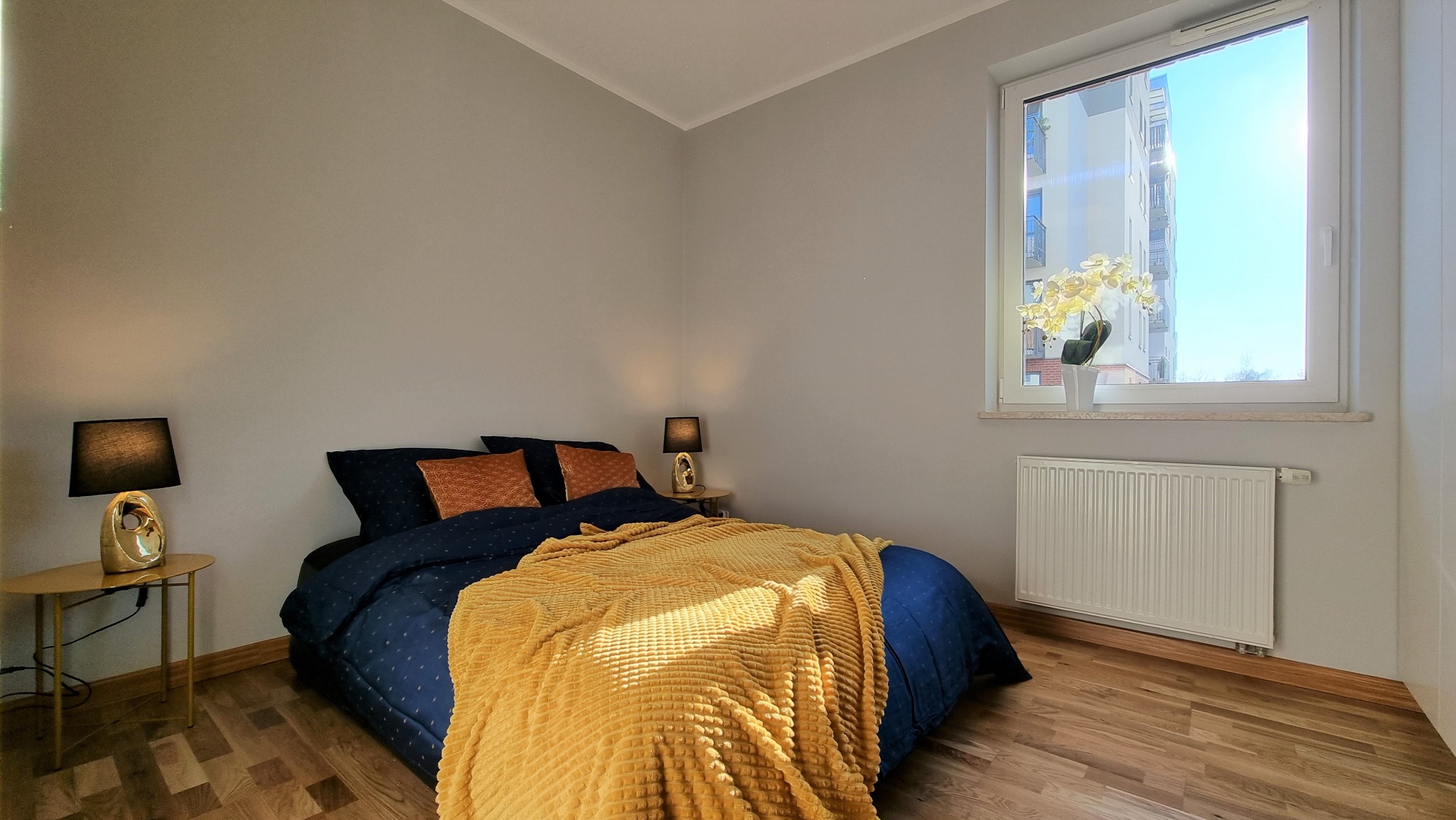 Jasne wnętrze sypialni z drewnianą podłogą, łóżkiem z niebieską pościelą i żółtym pledem. Na szafkach nocnych lampki, a na parapecie kwiaty. Widok z okna na budynek.