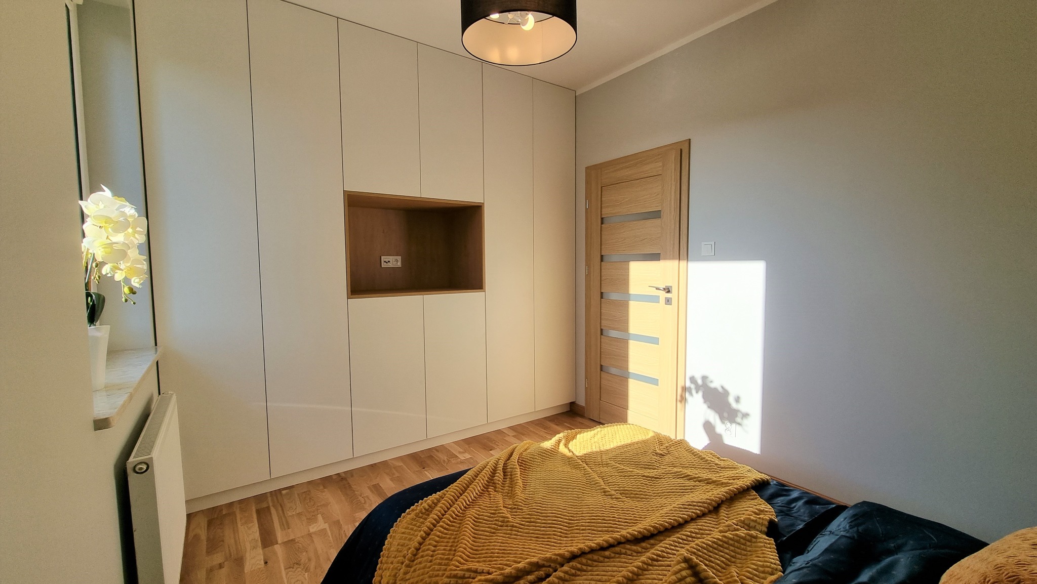 Jasne wnętrze sypialni z zabudowaną szafą wnękową z drewnianą wnęką, drewnianą podłogą i drzwiami. Na pierwszym planie łóżko z żółtą narzutą.