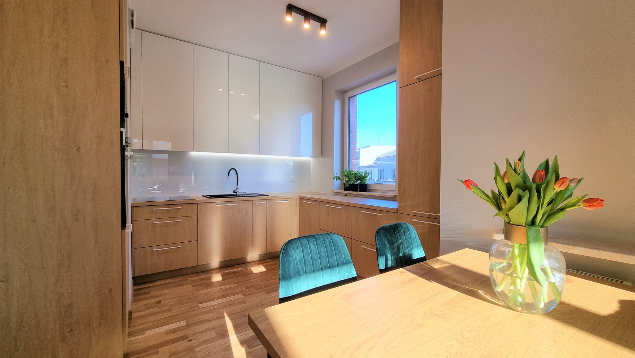 Jasna kuchnia z drewnianymi szafkami i białymi frontami, oświetlona listwą LED pod szafkami. Na stole wazon z tulipanami i dwa turkusowe krzesła.