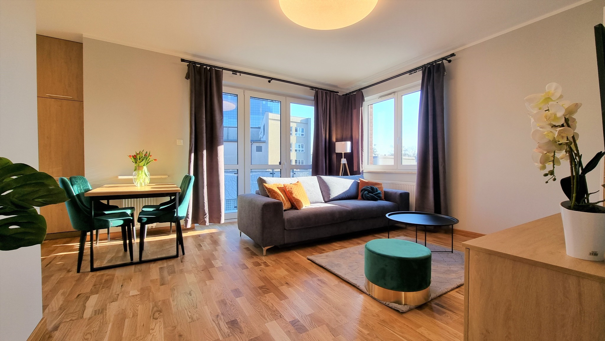 Jasne wnętrze salonu z drewnianą podłogą, szarą sofą z kolorowymi poduszkami, zielonymi krzesłami przy stole i dużymi oknami z zasłonami.