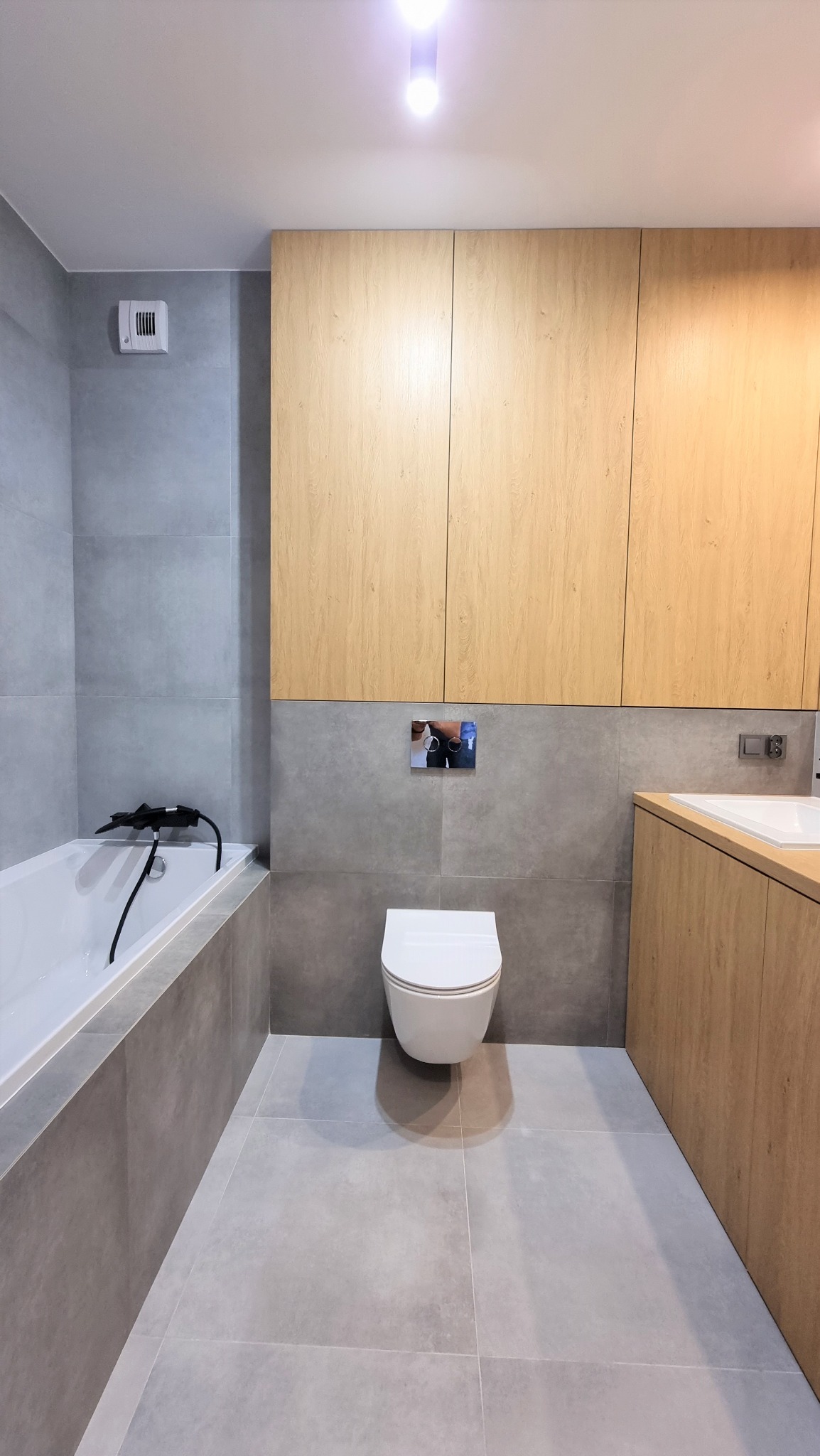 Nowoczesna łazienka z szarymi płytkami, drewnianymi szafkami i podwieszaną toaletą. Minimalistyczny design, oświetlenie punktowe, wanna z czarną armaturą.