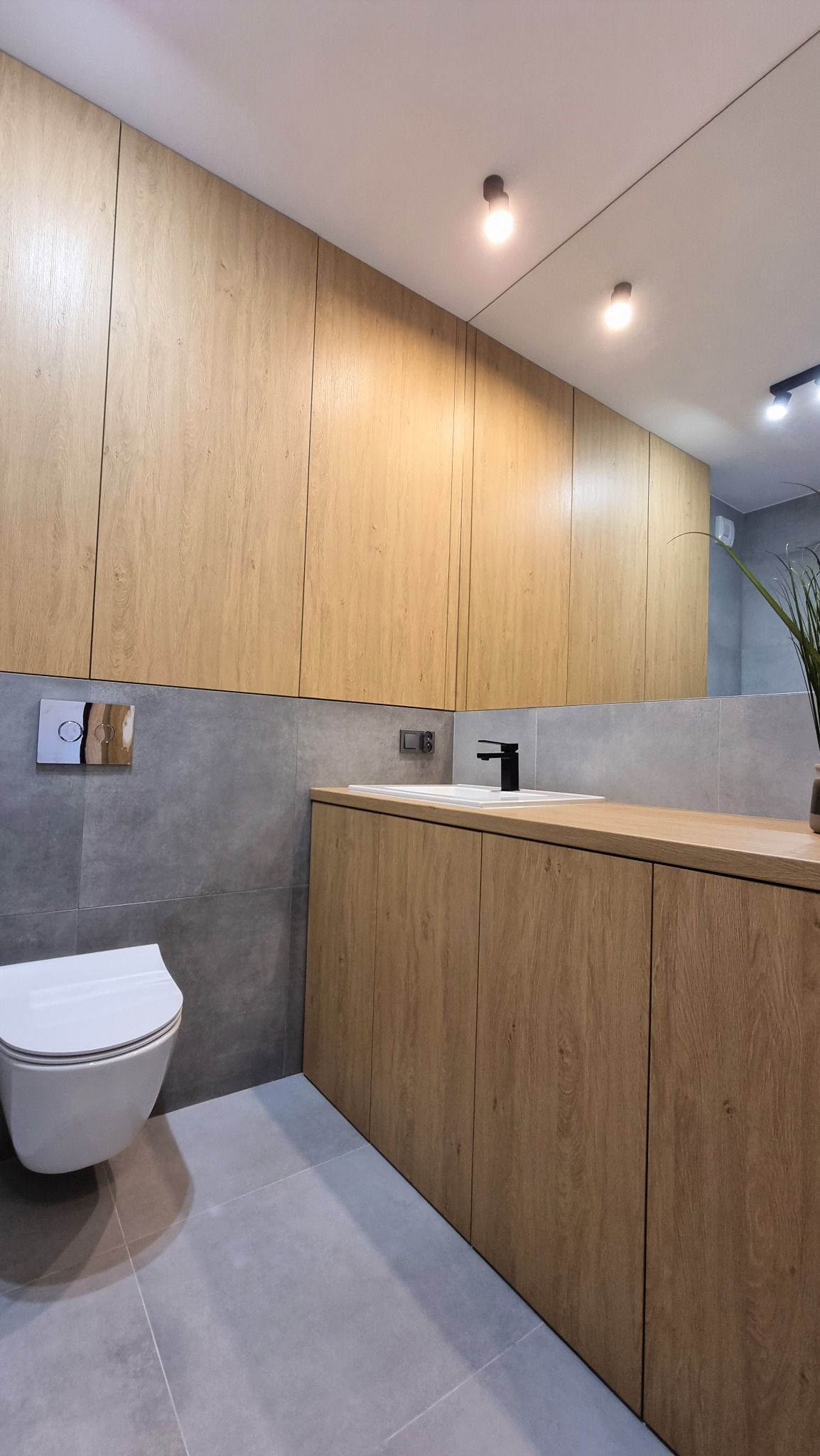 Nowoczesna łazienka po remoncie: szare płytki, drewniane szafki pod umywalką i wiszące nad nią, minimalistyczna armatura, podwieszana toaleta. Oświetlenie punktowe.