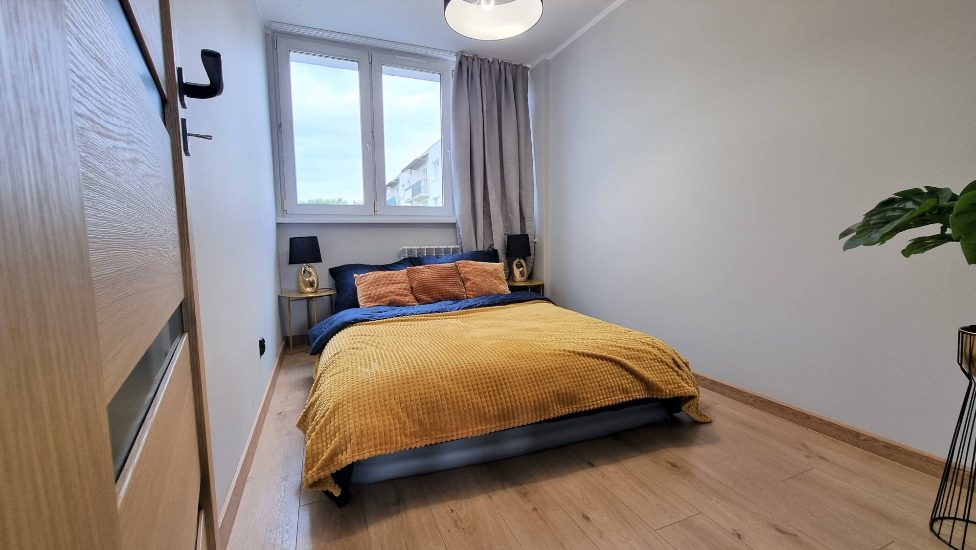 Jasna sypialnia z drewnianą podłogą i łóżkiem z żółtą narzutą. Widoczne okno z szarymi zasłonami, drzwi z klamką i roślina doniczkowa. Minimalistyczny wystrój.