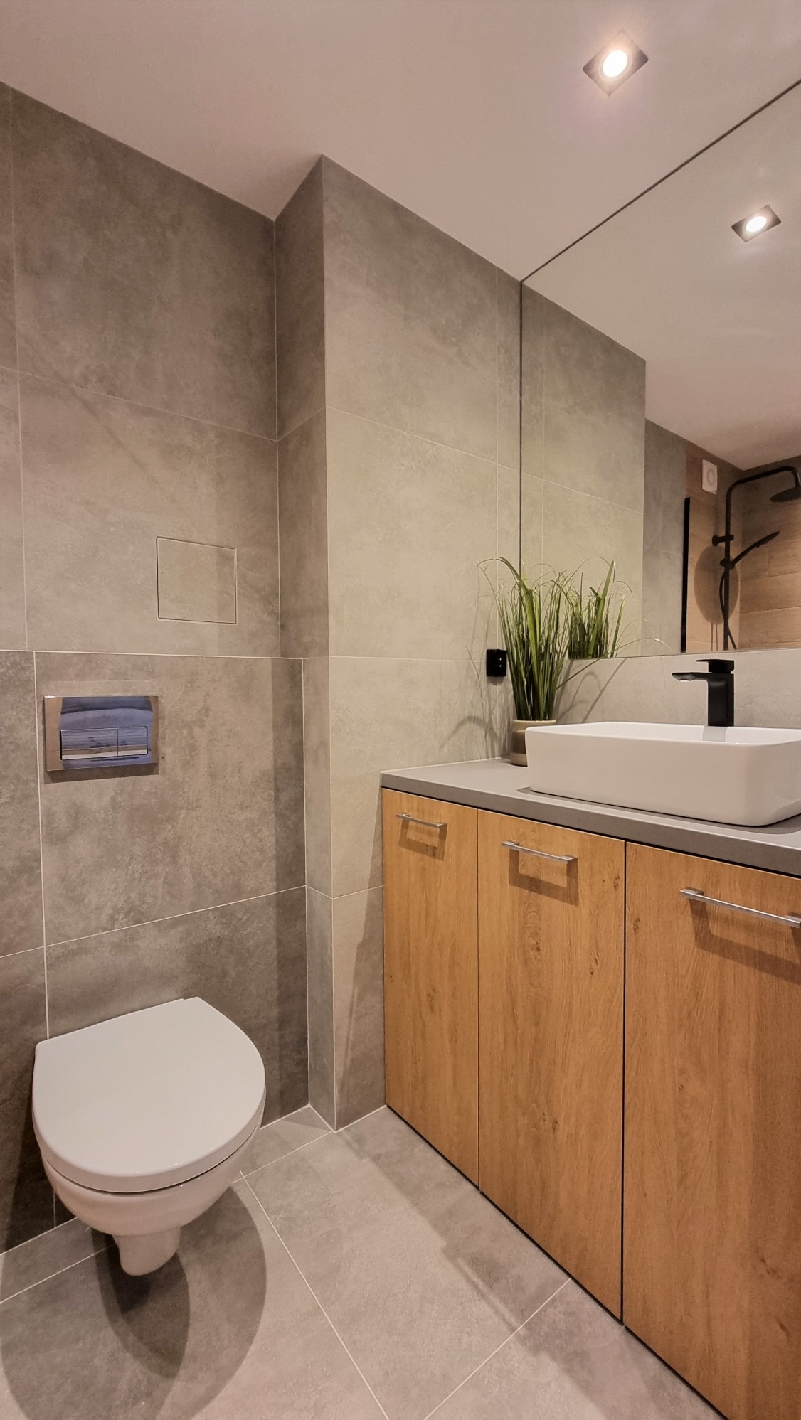 Nowoczesna łazienka z szarymi płytkami, drewnianą szafką pod umywalką i minimalistyczną armaturą. Uwagę zwraca duża powierzchnia płytek i ukryty rewizyjny właz.