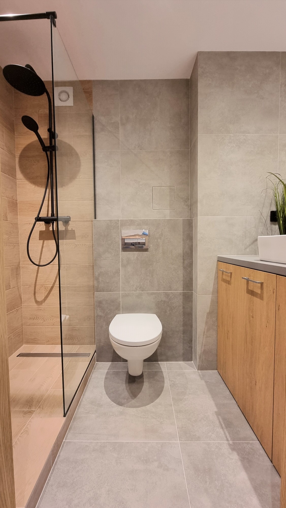 Nowoczesna łazienka z kabiną prysznicową typu walk-in, toaletą podwieszaną i szafką pod umywalką w odcieniach szarości i drewna. Minimalistyczny design, płytki wielkoformatowe.