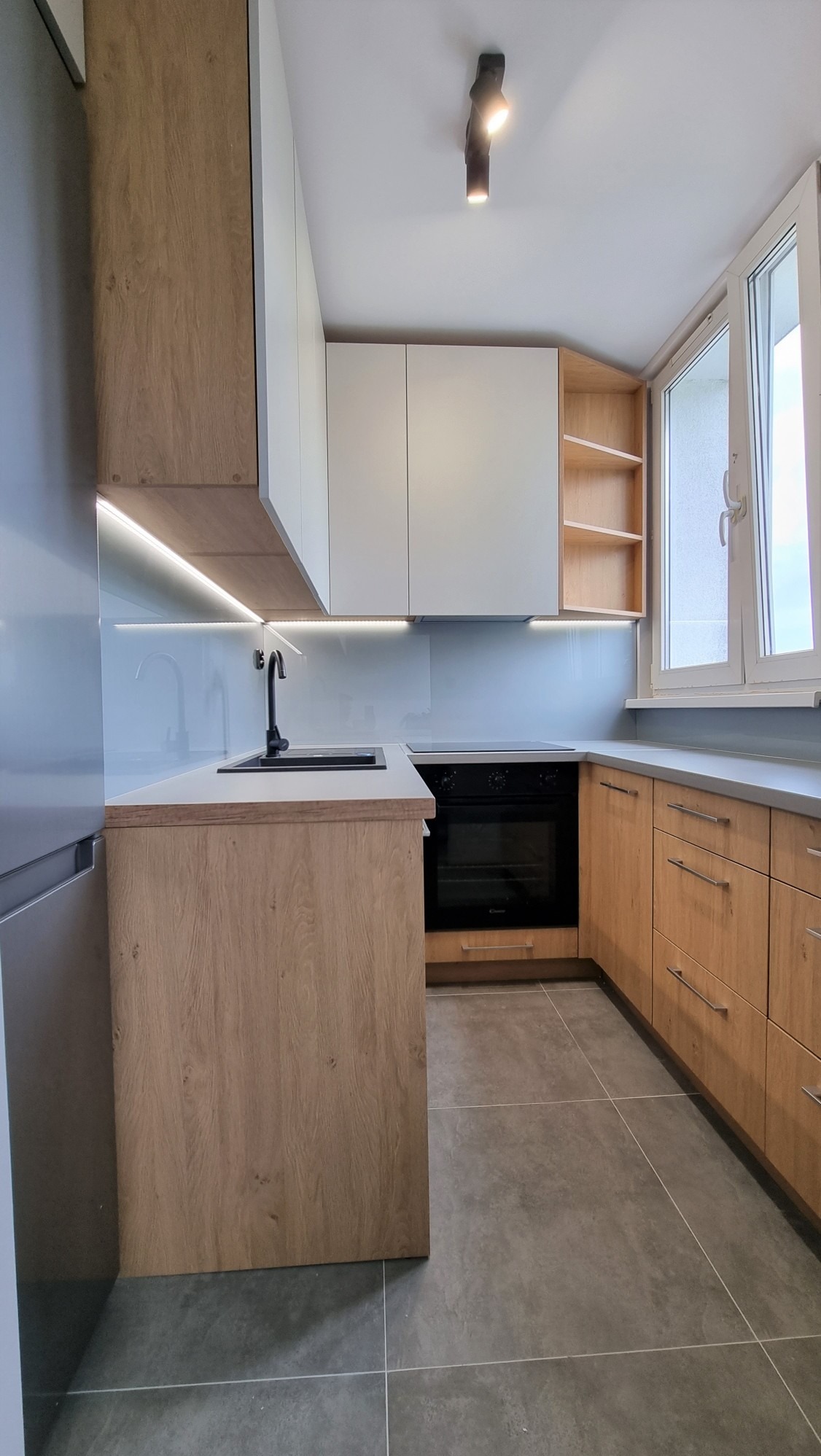 Nowoczesna kuchnia z drewnianymi i białymi szafkami, szarymi płytkami podłogowymi i oknem z widokiem na zewnątrz. Oświetlenie LED pod szafkami dodaje blasku.