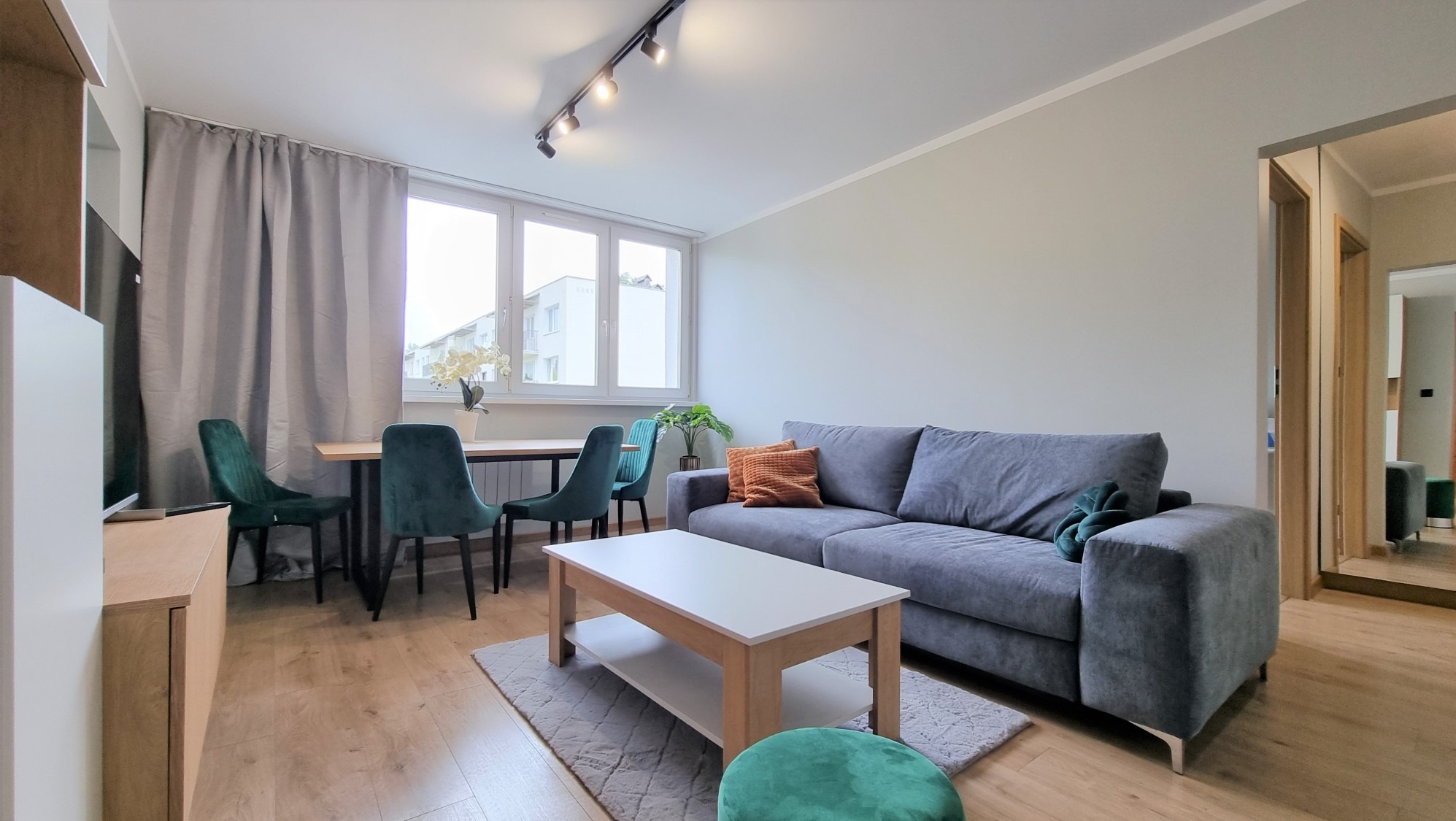 Jasne wnętrze salonu z szarą sofą, drewnianym stołem i zielonymi krzesłami. Parkiet na podłodze, okno z widokiem na zewnątrz. Nowoczesne oświetlenie sufitowe.