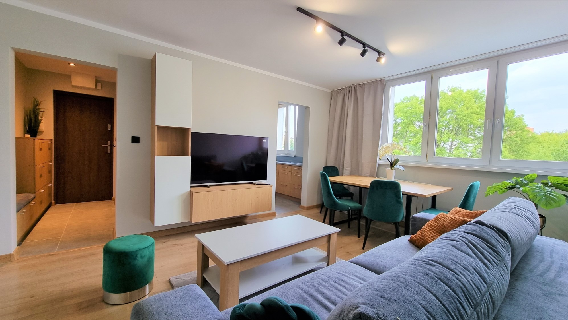 Jasne wnętrze salonu z drewnianą podłogą, szarą sofą i zielonymi akcentami. Widok na stół z krzesłami i okno z widokiem na zieleń. Nowoczesne oświetlenie sufitowe.