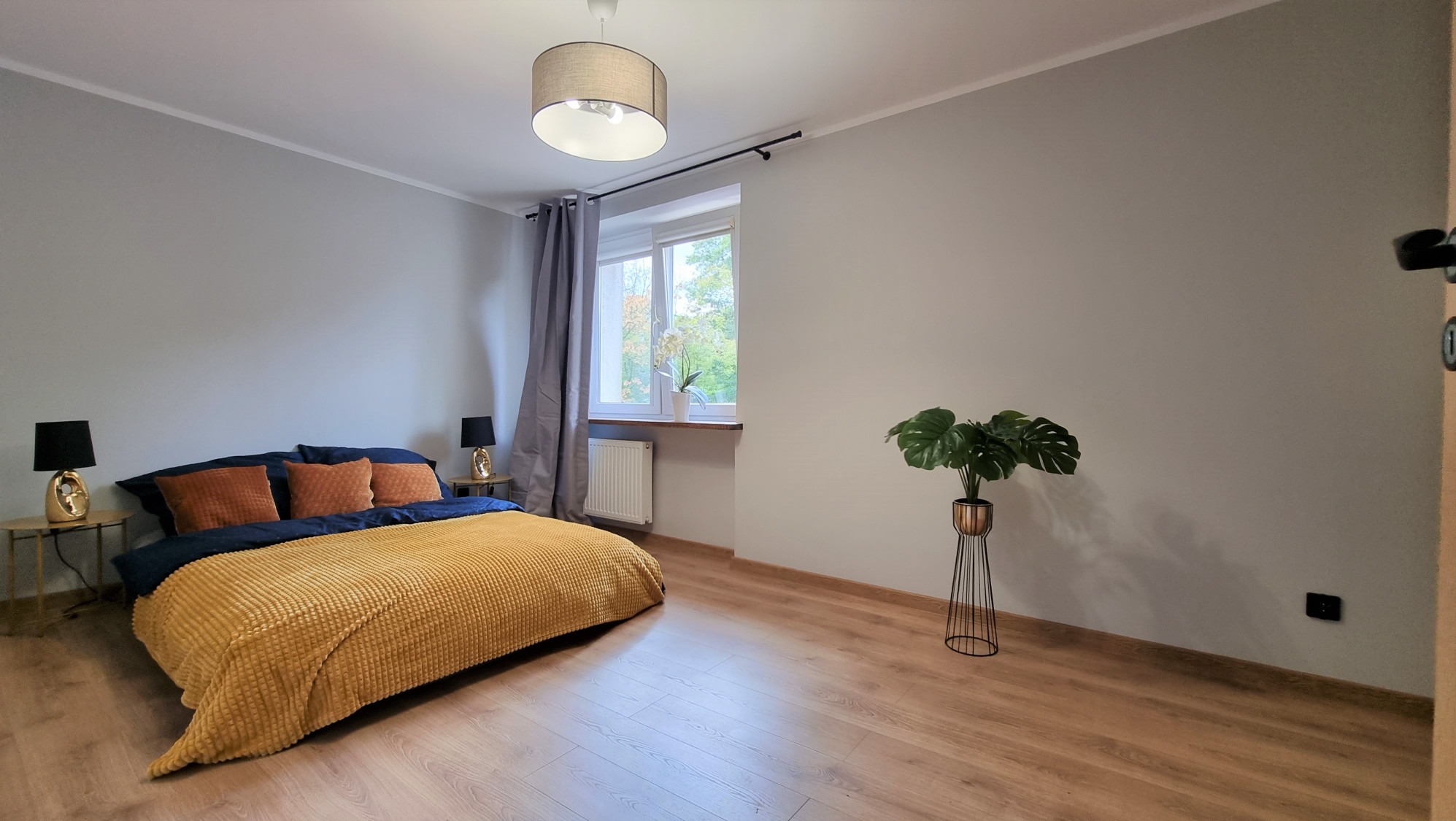 Jasna sypialnia z drewnianą podłogą, żółtą narzutą na łóżku, lampkami nocnymi i rośliną w ozdobnej donicy. Ściany w odcieniach szarości, okno z zasłonami.