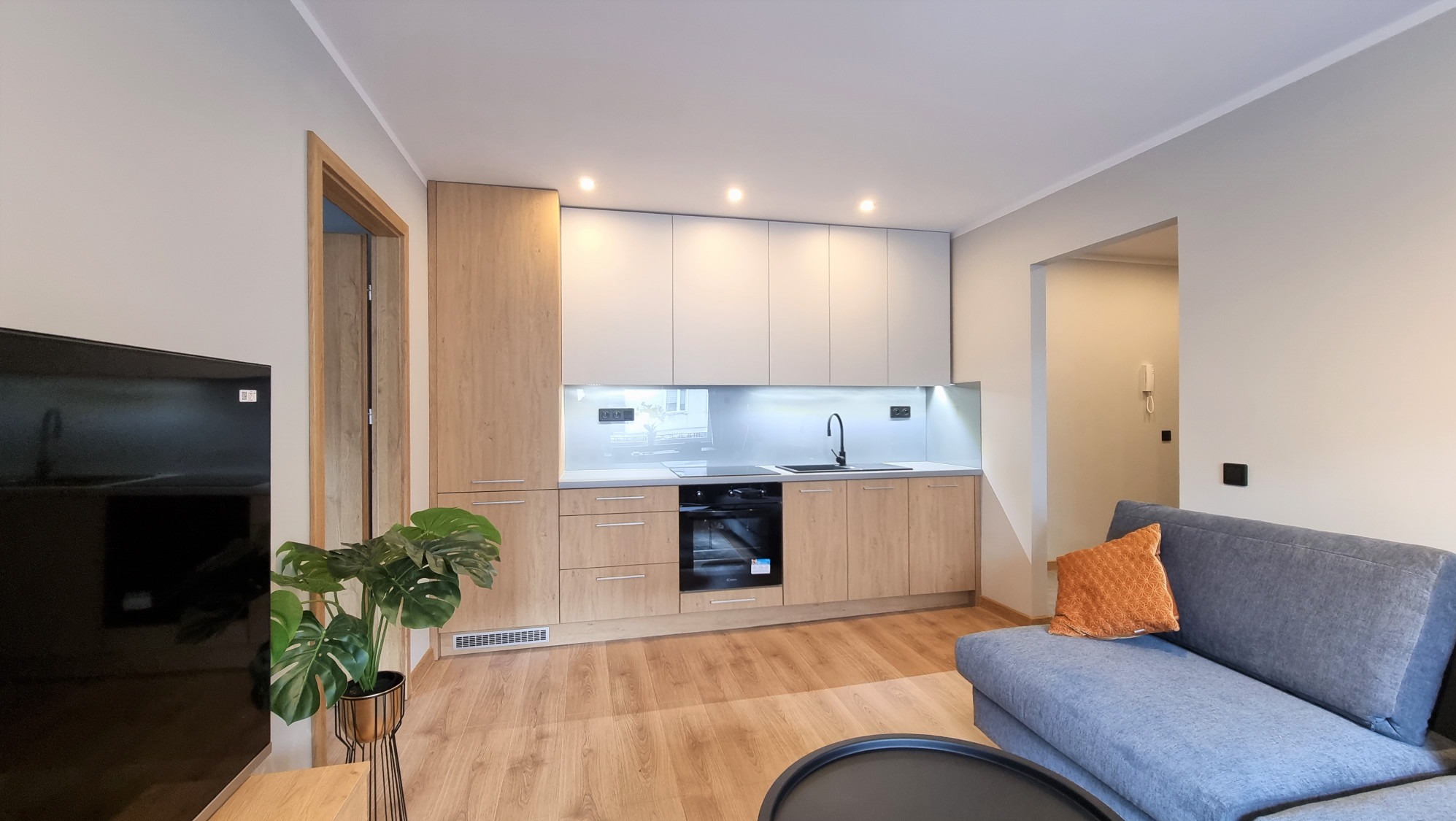 Nowoczesna kuchnia z drewnianymi frontami i szarymi blatami, oświetlona punktowo, widok z salonu z szarą sofą i rośliną doniczkową. Minimalistyczny design.