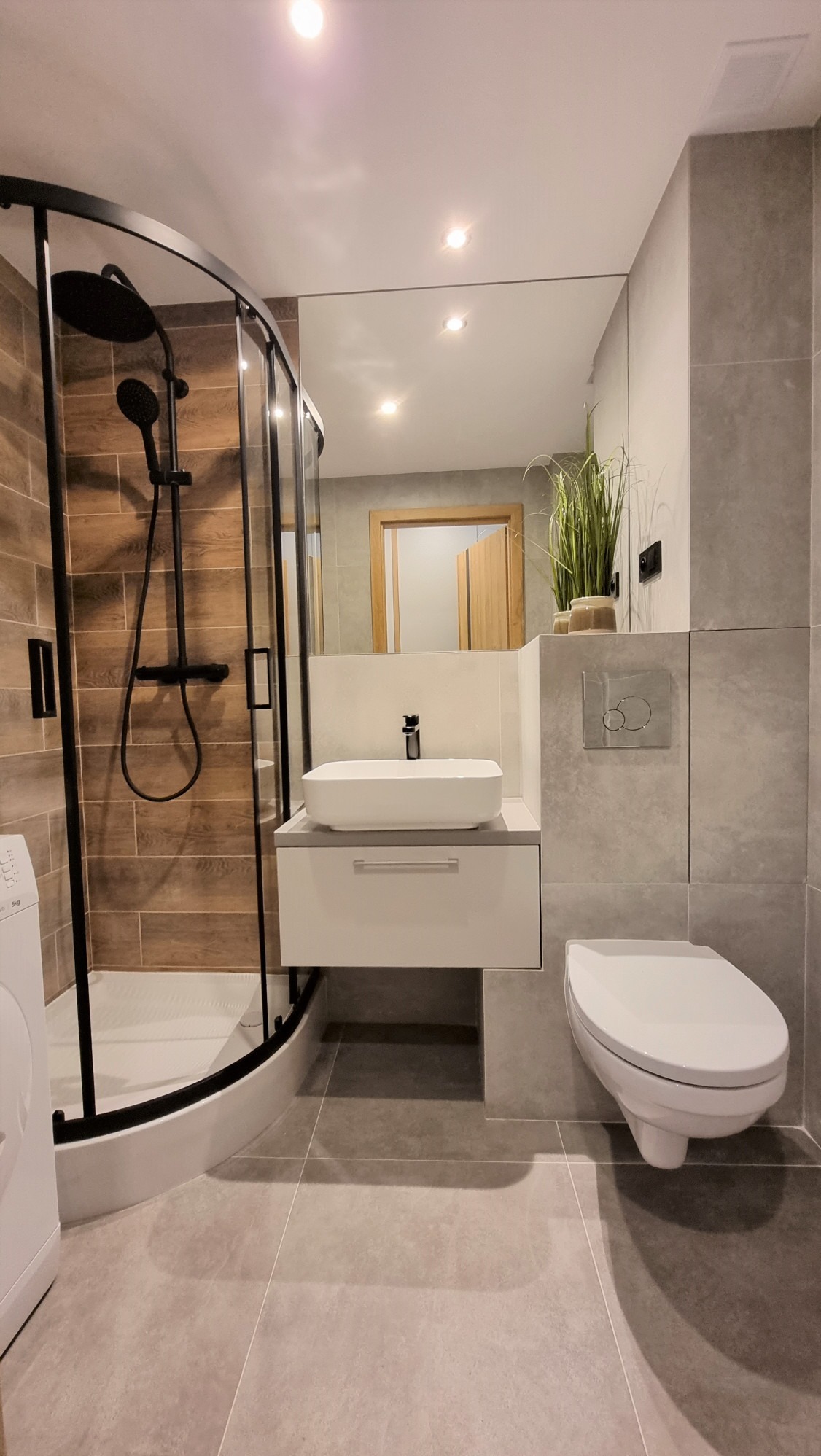 Nowoczesna łazienka z kabiną prysznicową typu walk-in, umywalką nablatową na szafce i podwieszaną toaletą. Uwagę zwracają brązowe płytki imitujące drewno w kabinie.