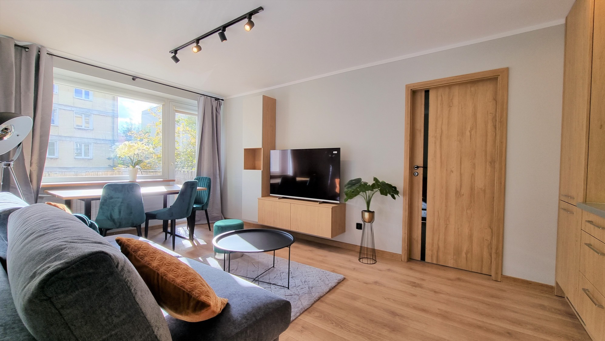 Jasne wnętrze salonu z drewnianą podłogą, szarą sofą i stołem jadalnym przy oknie. Widoczna zabudowa TV z szafkami i drzwi wewnętrzne z czarnym akcentem.