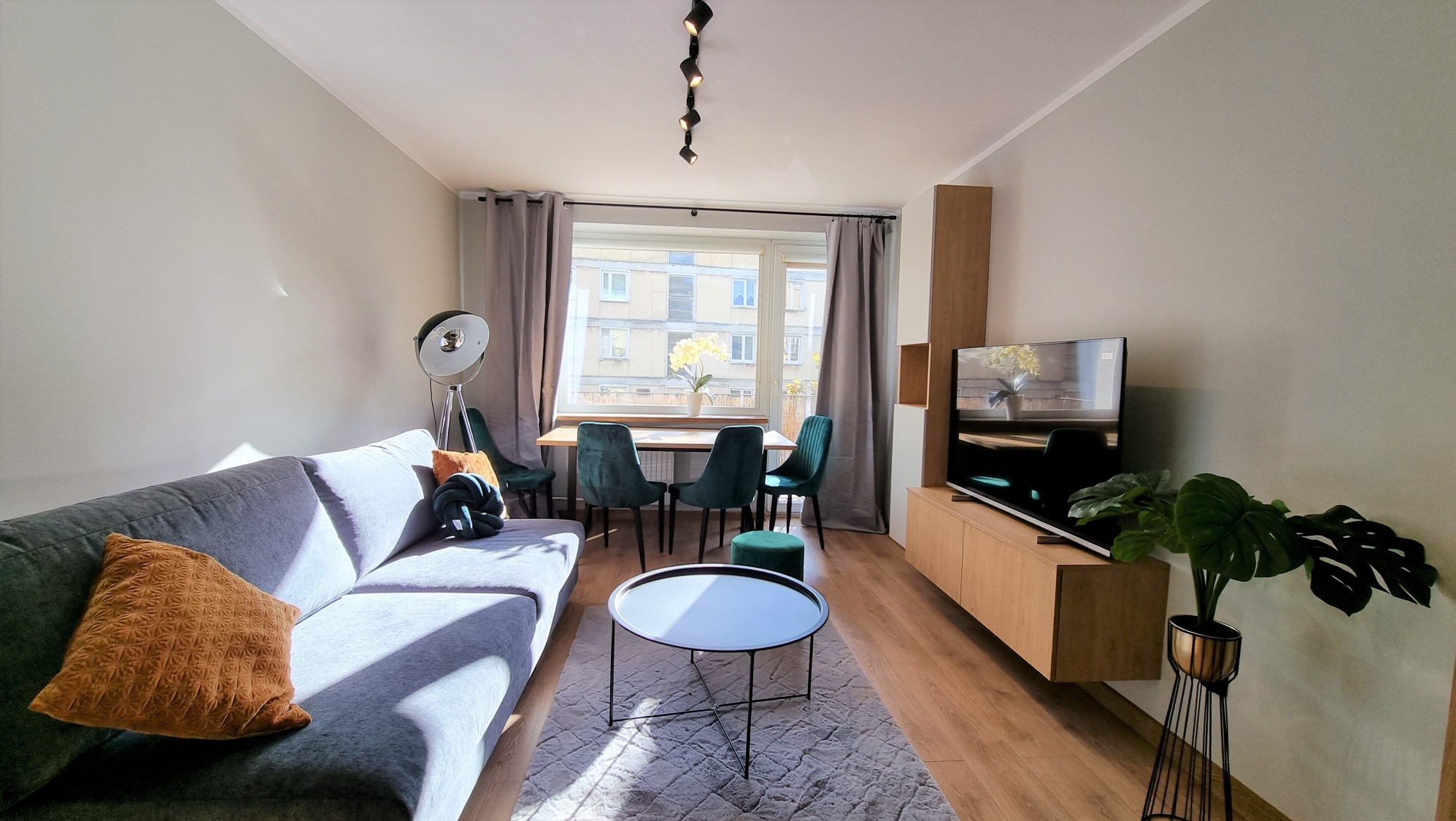 Jasne wnętrze salonu z szarą sofą, okrągłym stolikiem kawowym i drewnianą szafką RTV. W tle stół z krzesłami i widok na budynek przez okno.