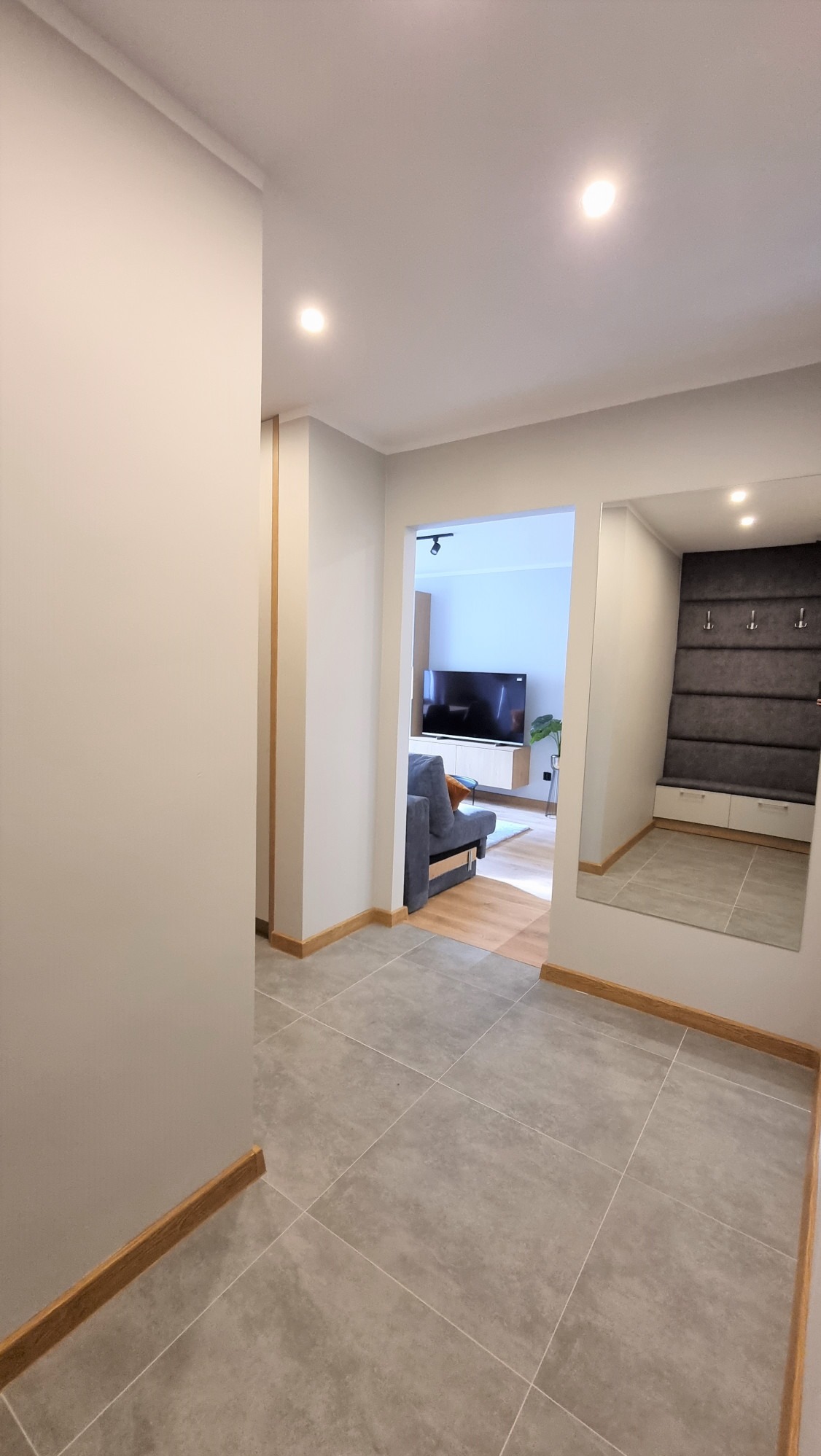 Nowoczesny przedpokój z szarą podłogą z płytek, lustrem i widokiem na salon. Neutralna kolorystyka ścian i drewniane listwy przypodłogowe. Minimalistyczny design.