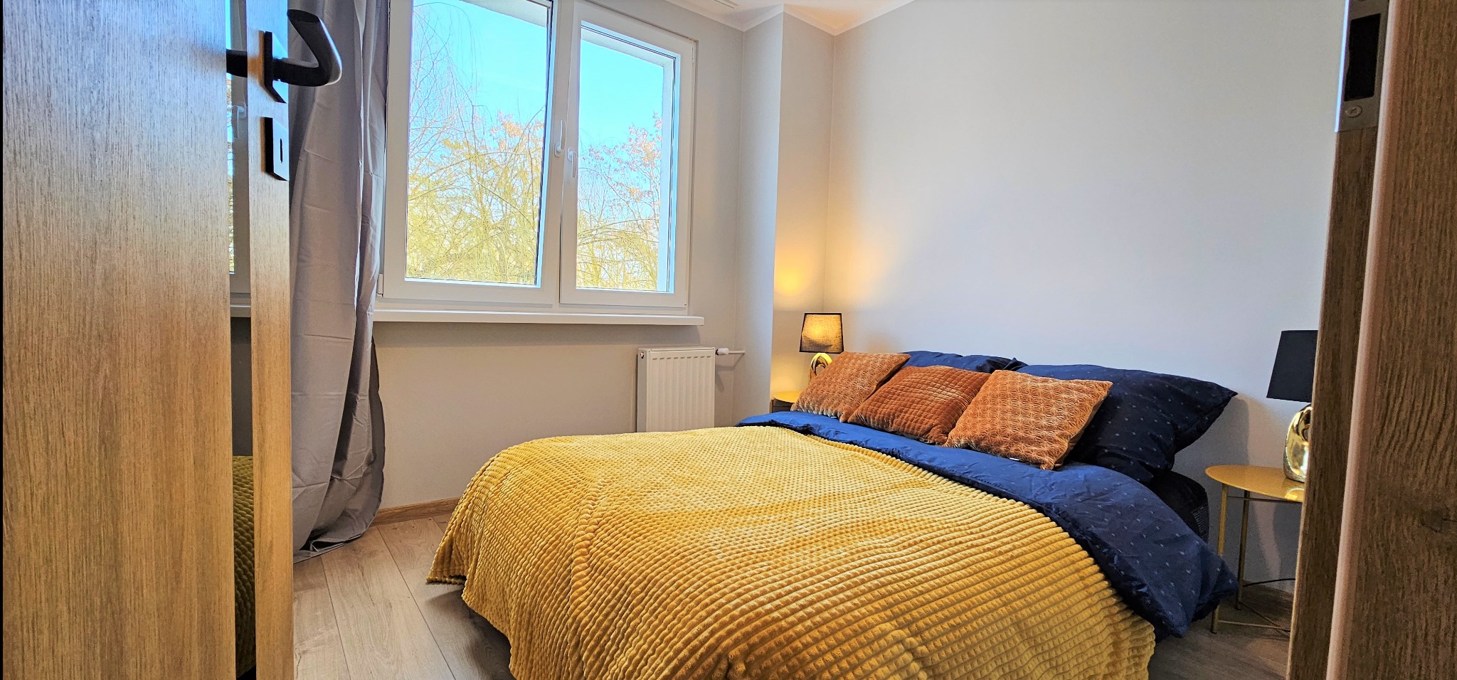 Jasna sypialnia z łóżkiem w odcieniach żółci i granatu, widok przez uchylone drzwi z drewnianą okleiną. Okno z widokiem na drzewa w tle.