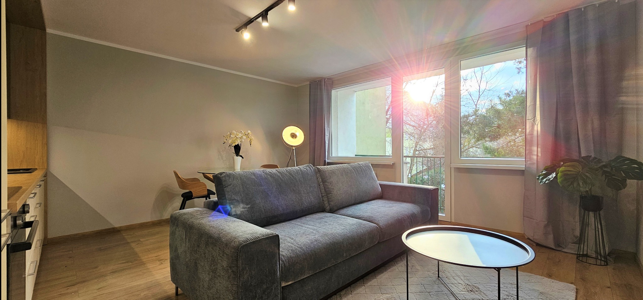 Jasne wnętrze salonu po remoncie: szara sofa, okrągły stolik kawowy, drewniana podłoga, duże okna z widokiem na zieleń, lampa podłogowa i oświetlenie sufitowe.