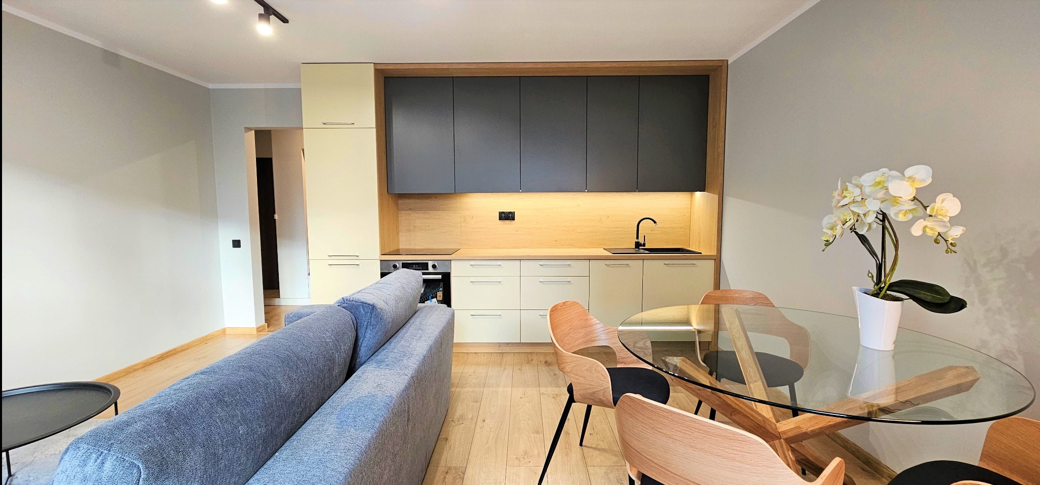 Nowoczesna kuchnia z szarymi szafkami i drewnianym blatem, oświetlona punktowo, z okrągłym stołem i sofą w tle. Minimalistyczny design.