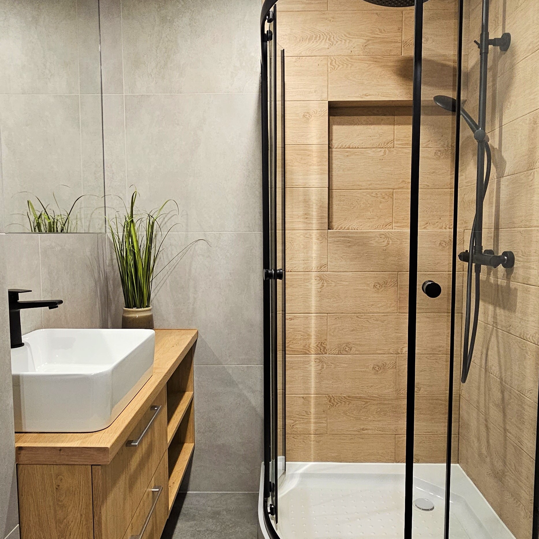 Nowoczesna łazienka z kabiną prysznicową typu walk-in, umywalką nablatową na drewnianej szafce i szarymi płytkami. Minimalistyczny design z akcentami drewna.