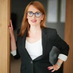 Kobieta w okularach i czarnej marynarce pozuje w drzwiach, z zegarkiem na ręce i naszyjnikiem z sercem.