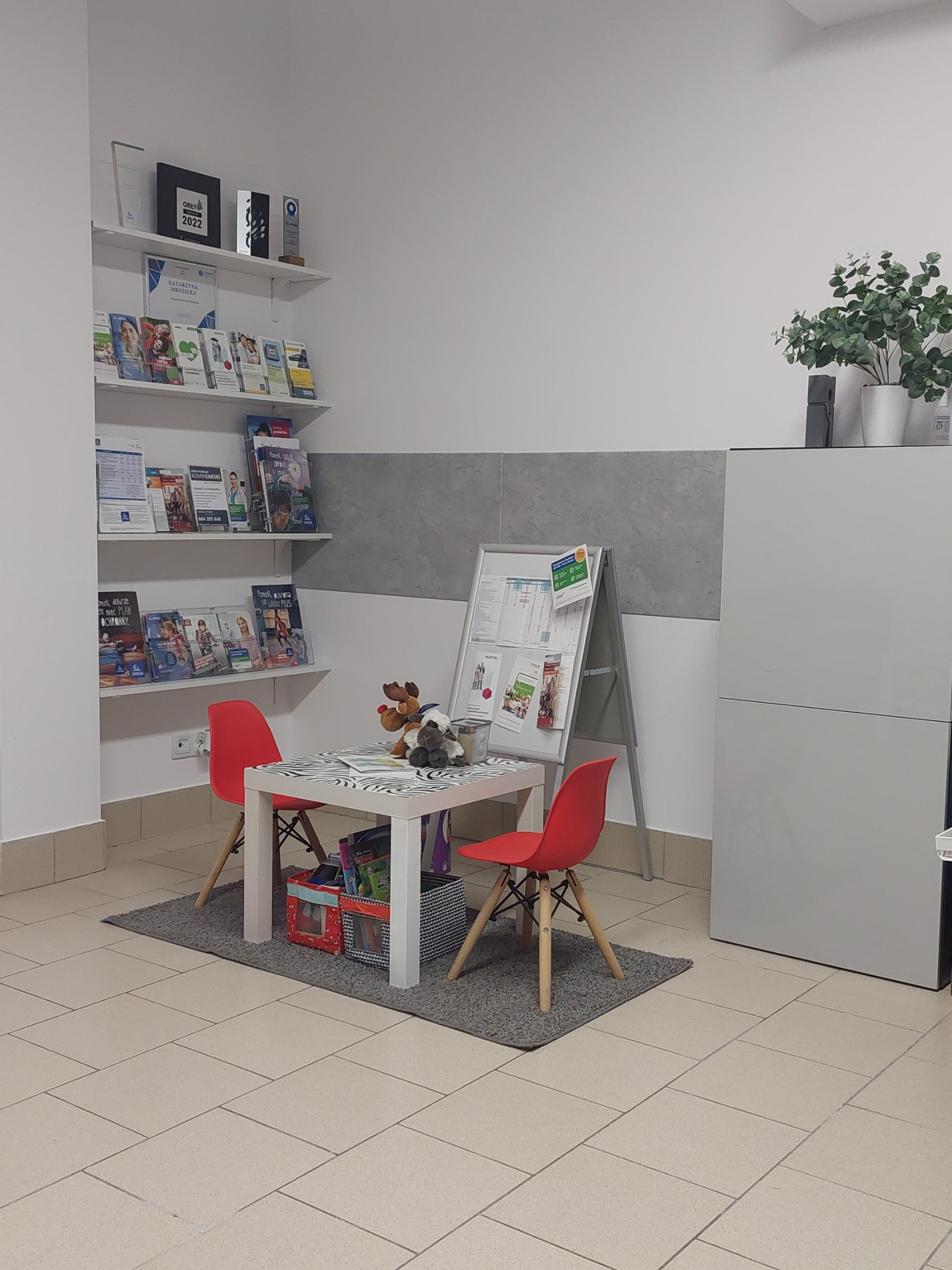 Jasne wnętrze biura z kącikiem dla dzieci: stolik z dwoma czerwonymi krzesłami, zabawki, regał z broszurami i dyplomami na ścianie, tablica informacyjna na sztalugach.