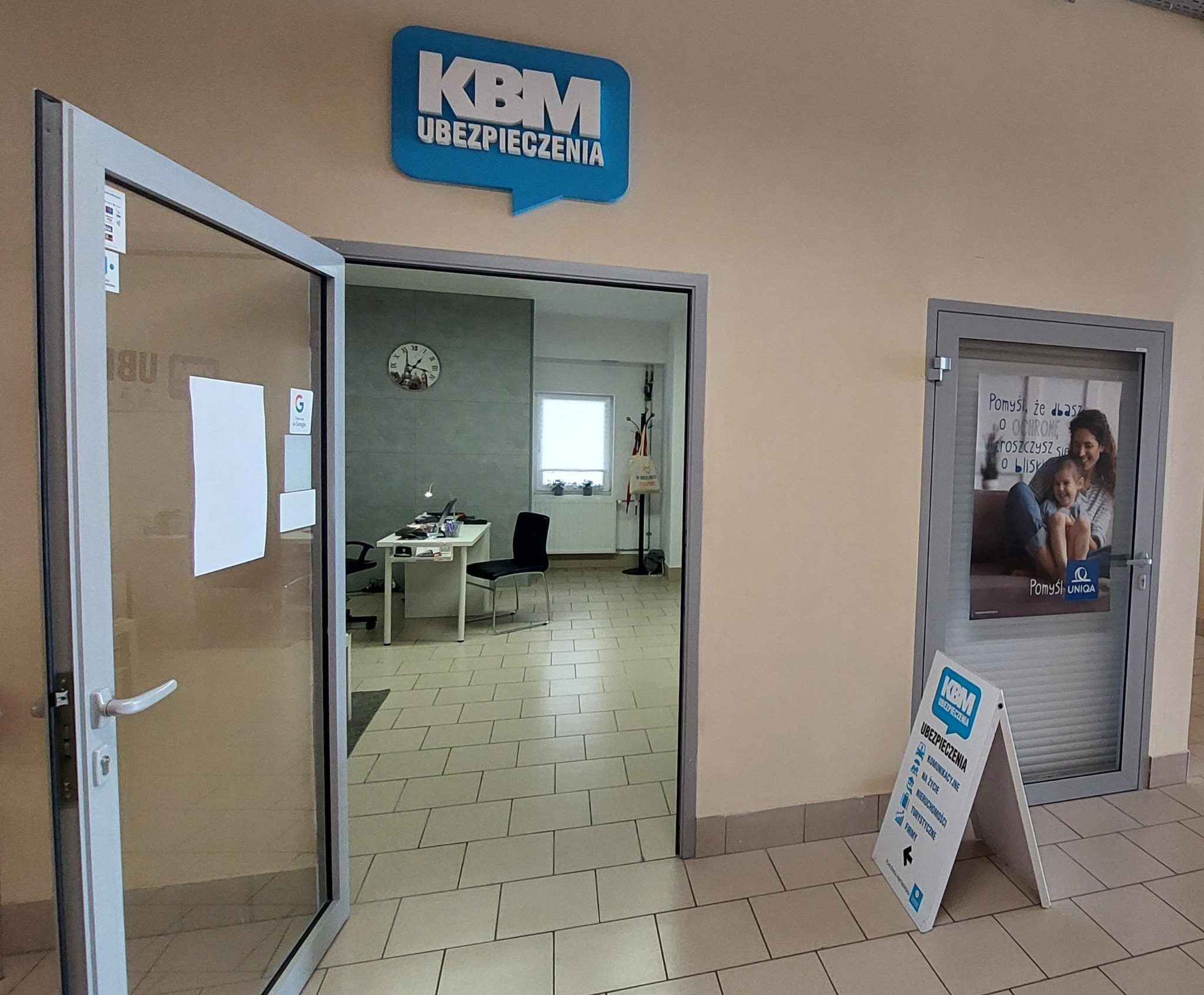 Wnętrze biura agencji ubezpieczeniowej KBM z otwartymi drzwiami wejściowymi, widoczny znak firmowy nad wejściem i tablica reklamowa z ofertą ubezpieczeń na życie, komunikacyjnych, nieruchomości...