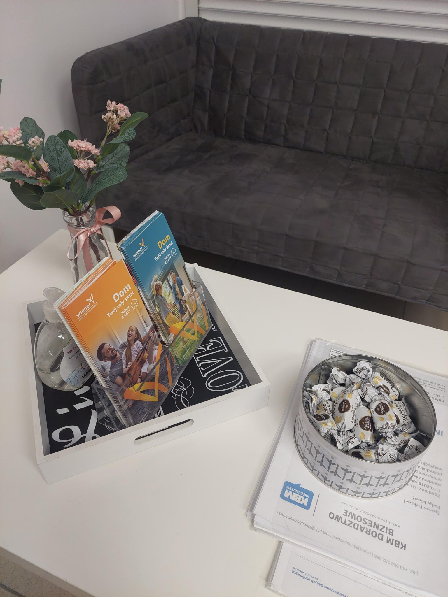 Stolik w biurze z ulotkami Wiener Insurance Group, żel antybakteryjny, puszka cukierków Wawel i dokumenty KBM Doradztwo Biznesowe