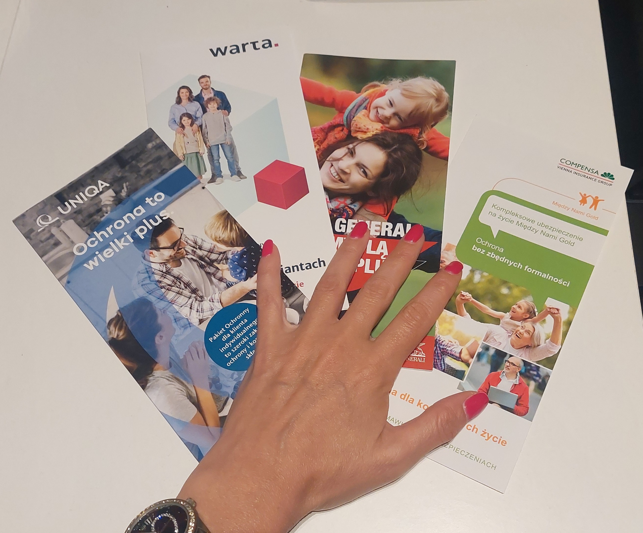 Rozłożone ulotki reklamowe różnych firm ubezpieczeniowych, przytrzymywane dłonią z czerwonym manicure, na białym tle biurka.