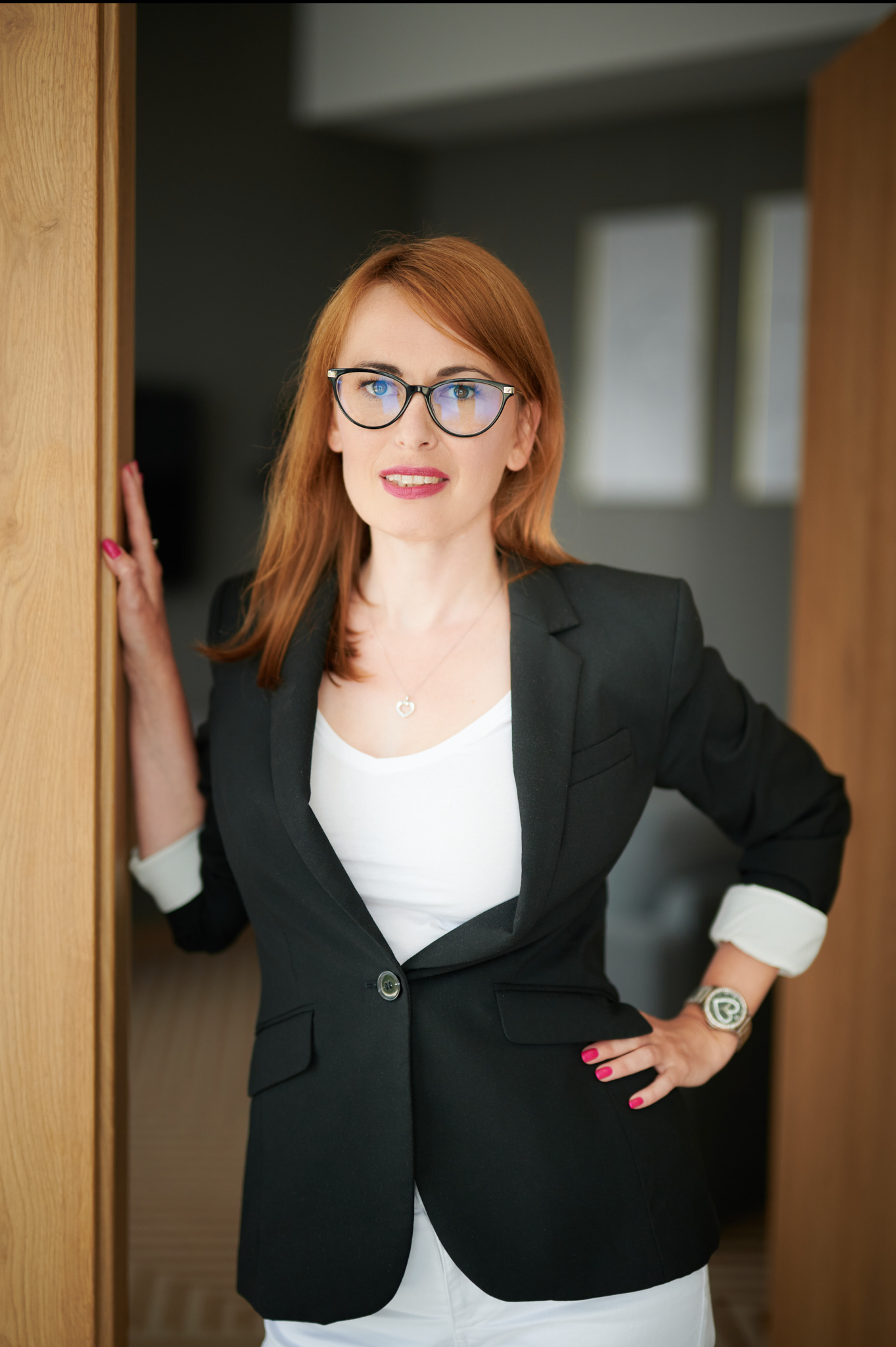 Kobieta w okularach i czarnej marynarce pozuje w drzwiach, z zegarkiem na ręce i naszyjnikiem z sercem.
