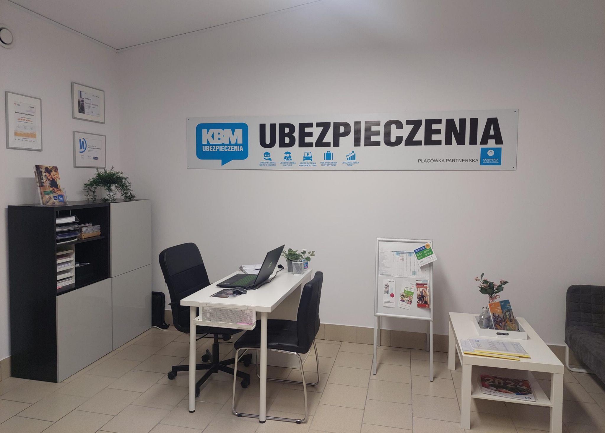 Wnętrze biura agencji ubezpieczeniowej z biurkiem, komputerem, krzesłami, tablicą informacyjną i regałem na dokumenty. Na ścianie widoczny baner reklamowy z logo firmy i zakresem usług.
