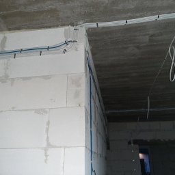 Łukasz Mika - Białe kable elektryczne ułożone wzdłuż surowych ścian z betonu komórkowego, zabezpieczone czarnymi uchwytami. Fragment instalacji w nowym budynku.