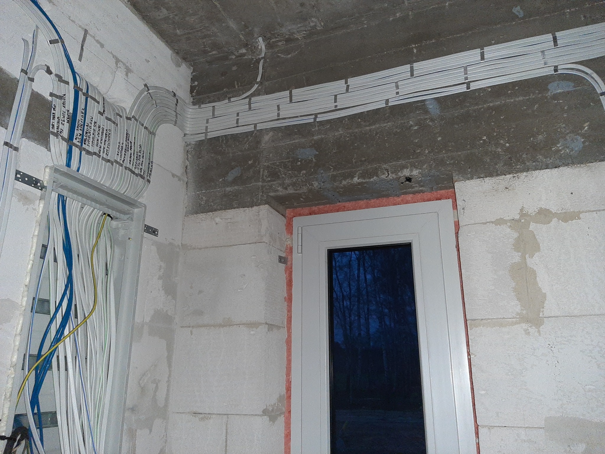 Białe korytka kablowe ułożone na suficie i ścianie w surowym wnętrzu. Widoczne przewody w peszlach przy futrynie drzwiowej. Ściany z betonu komórkowego.