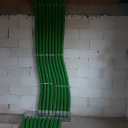 Równolegle ułożone, zielone rury wentylacyjne z tworzywa sztucznego, przymocowane metalowymi obejmami do ściany z białych bloczków betonowych w surowym wnętrzu budynku.