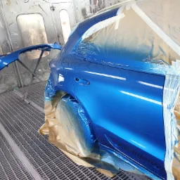 Niebieski samochód w kabinie lakierniczej, częściowo zamaskowany folią i taśmą, zderzak obok na stojaku.