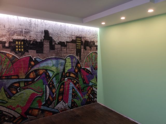 Wnętrze pokoju z jedną ścianą pomalowaną na jasnozielono i drugą pokrytą fototapetą z motywem graffiti przedstawiającym abstrakcyjny krajobraz miejski w odcieniach szarości, czerni, czerwieni...