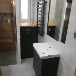 Nowoczesna łazienka z szarą armaturą, umywalką nablatową na szafce z szufladami, grzejnikiem drabinkowym i szafką pod oknem z drewnianym blatem.