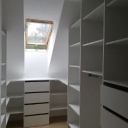 Biała garderoba z szufladami i półkami w pomieszczeniu ze skosem i oknem dachowym.