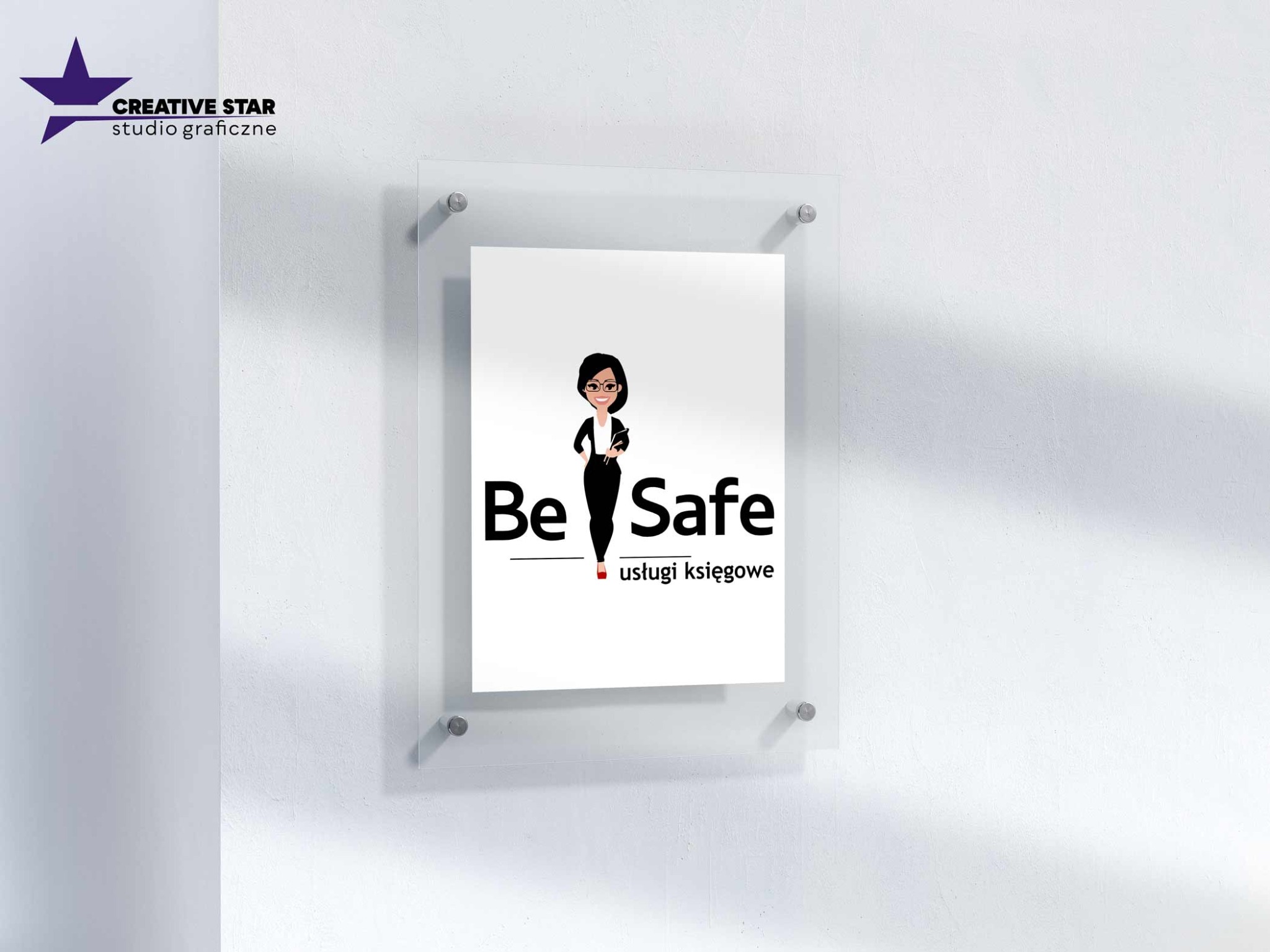 Tabliczka z logo 'Be Safe usługi księgowe' w przezroczystej ramce, przymocowana do białej ściany. W lewym górnym rogu logo 'Creative Star studio graficzne'.
