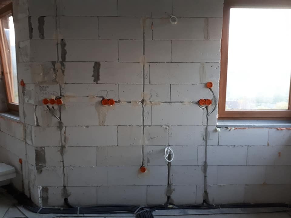 Surowa ściana z betonu komórkowego z widocznymi przewodami elektrycznymi w peszlach oraz puszkami instalacyjnymi z pomarańczowymi zabezpieczeniami, przygotowana do dalszych prac wykończeniowych.