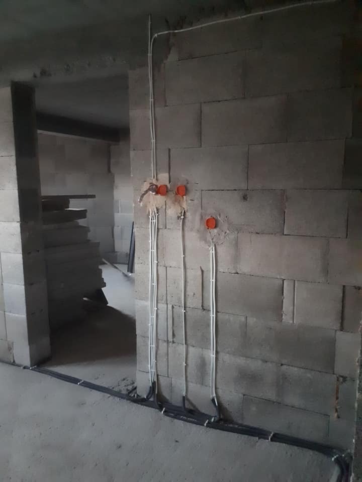 Surowy stan instalacji elektrycznej w budynku z pustaków: widoczne kable w peszlach na podłodze, białe przewody pionowo na ścianie oraz pomarańczowe puszki instalacyjne.