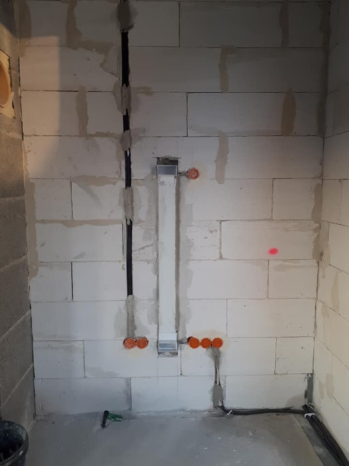 Ściana z betonu komórkowego w trakcie montażu instalacji elektrycznej: pionowe kable w bruzdach, puszki pod gniazdka i rura instalacyjna z metalowym elementem. Widoczny fragment podłogi i wiadro.