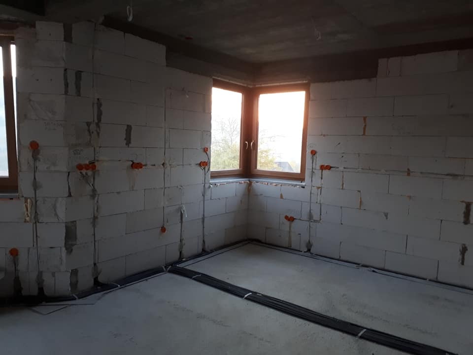 Surowy stan wnętrza z widoczną instalacją elektryczną: kable w peszlach prowadzone po ścianach z betonu komórkowego, pomarańczowe puszki instalacyjne, okna z widokiem na zewnątrz.