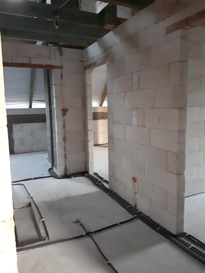 Surowy stan budynku z widocznymi przewodami elektrycznymi ułożonymi na podłodze i przymocowanymi do ściany, instalacja w trakcie realizacji.