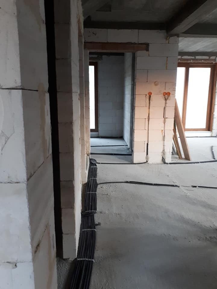 Wiązka kabli elektrycznych ułożona wzdłuż ściany z betonu komórkowego w surowym wnętrzu budynku w trakcie budowy, widoczne puszki instalacyjne na ścianie, w tle otwory drzwiowe.