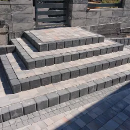 Schody z kostki brukowej prowadzące do furtki w ogrodzeniu, wykończone obrzeżami z drobnej kostki, z widocznym fragmentem muru oporowego w tle.