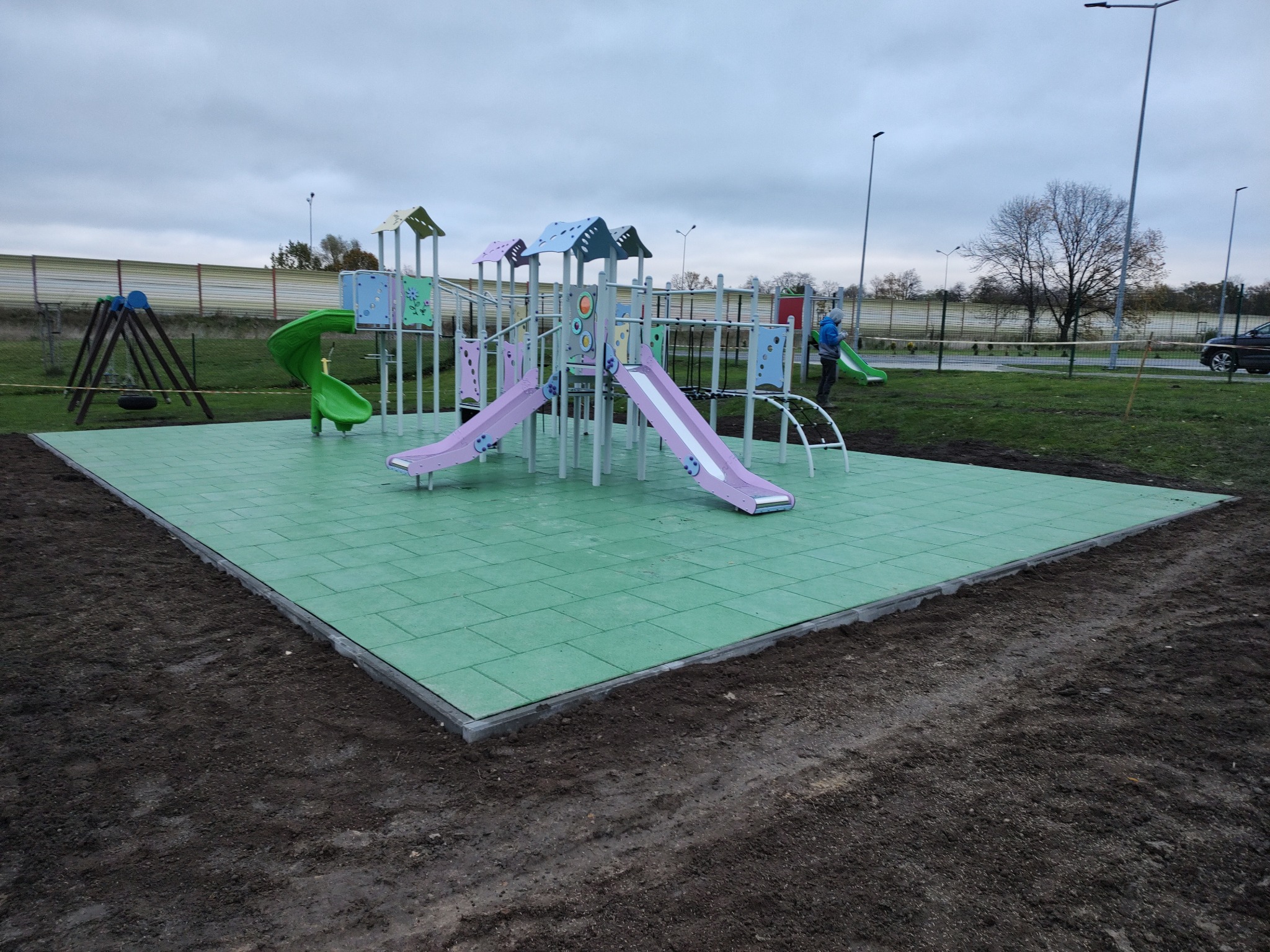 Nowo wybudowany plac zabaw z zieloną, gumową nawierzchnią, kolorowymi zjeżdżalniami i huśtawką na tle pochmurnego nieba i drzew bez liści.