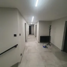 Pomalowany korytarz w neutralnych barwach z widocznym wiadrem farby, grzejnikiem i wieszakiem na ubrania.