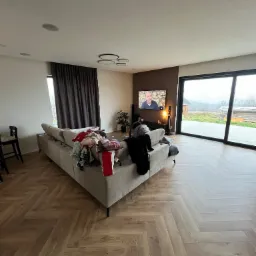 Salon z drewnianą podłogą ułożoną w jodełkę, beżowa sofa z ubraniami i prezentami, duże okno z widokiem na zewnątrz, telewizor na brązowej ścianie.